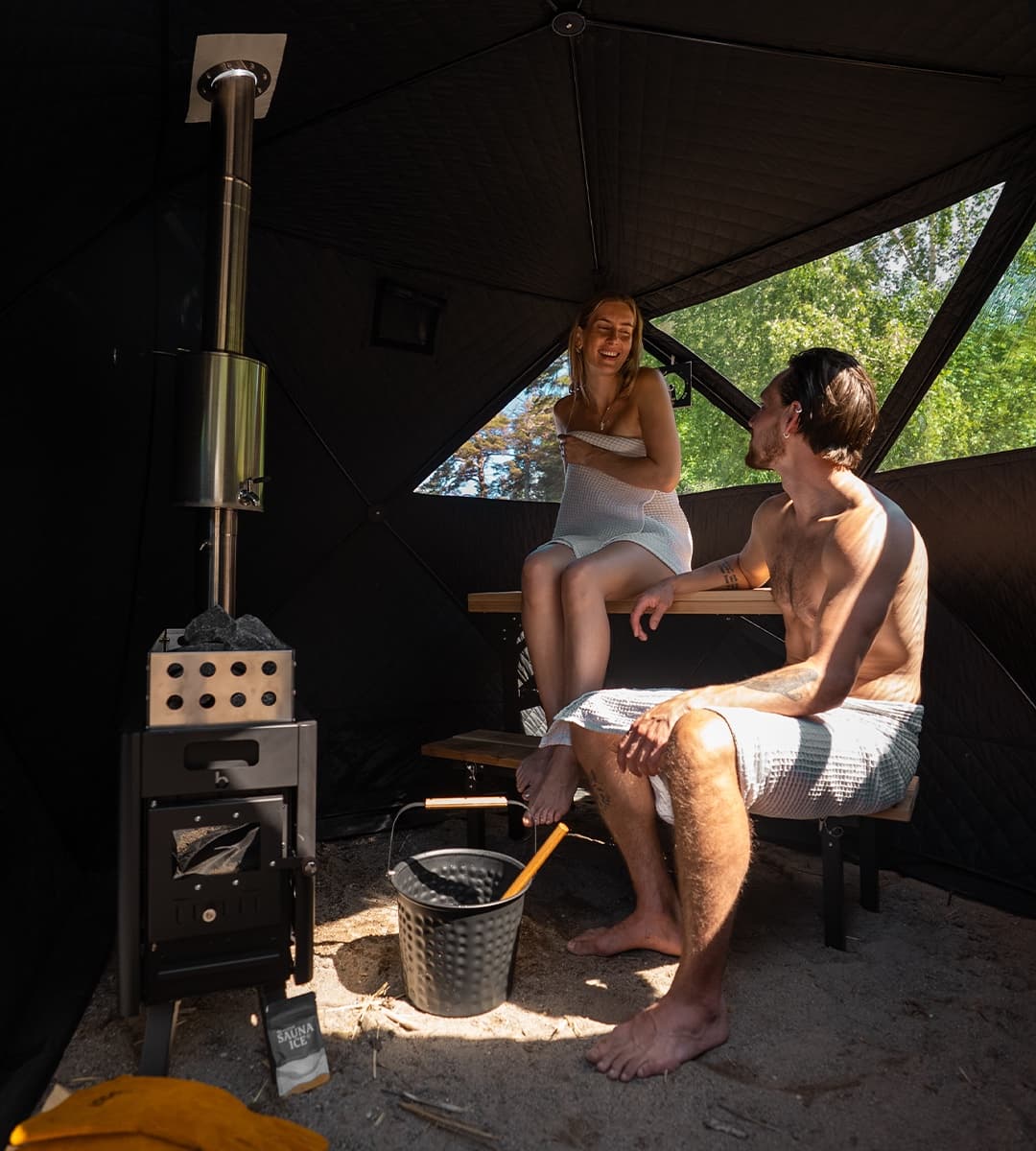 Inside the Näver Deluxe: two people sitting by the wood stove with a window open to the forest outside