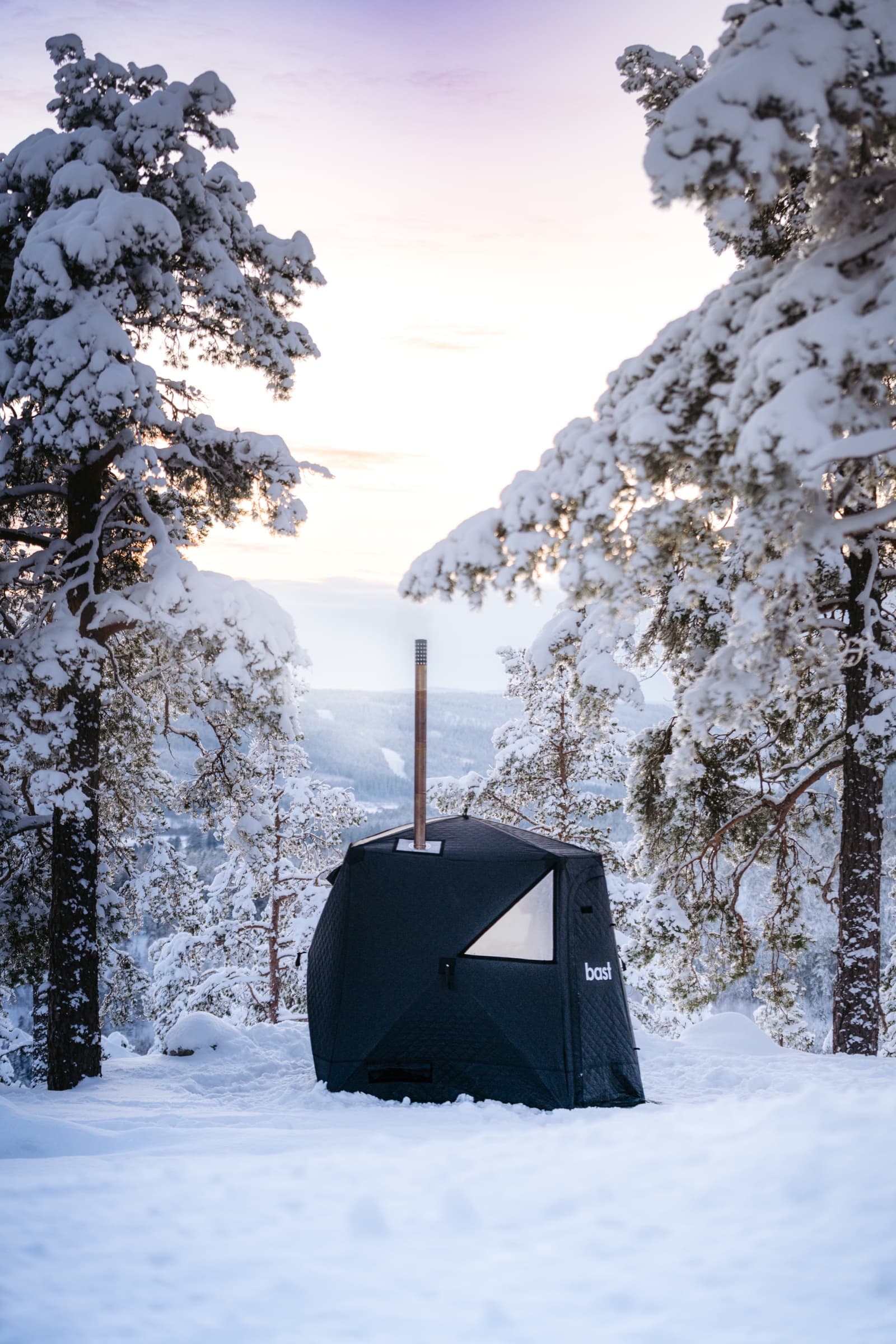 The Näver sauna tent in a snow-covered Nordic pine forest with a dusk sky above the valley